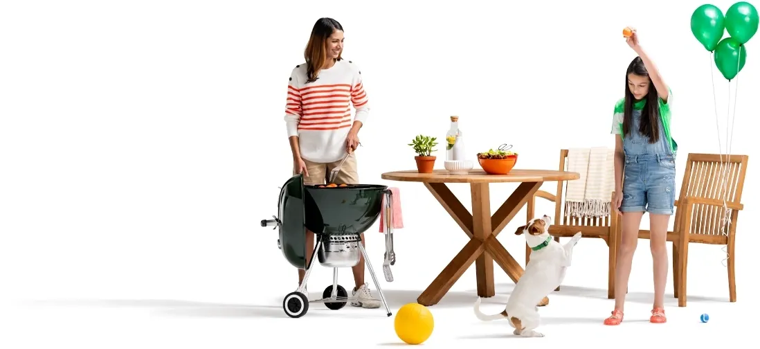 As mom tends the grill next to the outdoor patio furniture, her daughter plays fetch with their Jack Russell terrier A trio of green balloons are tied to the wooden picnic chair.